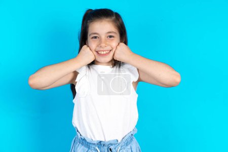 Photo for Happy kid keeps fists on cheeks smiles broadly and has positive expression being in good mood. Beautiful caucasian little girl posing over blue studio background - Royalty Free Image
