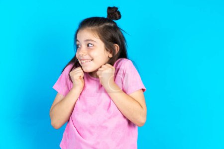 Photo for Kid clenches fists and awaits for something nice to happen. Beautiful caucasian little girl posing over blue studio background - Royalty Free Image