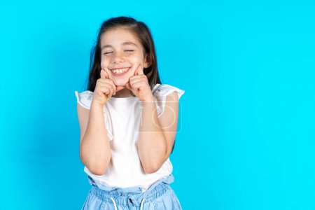 Photo for Pleased kid with closed eyes keeps hands near cheeks and smiles tenderly, imagines something very pleasant. Beautiful caucasian little girl posing over blue studio background - Royalty Free Image