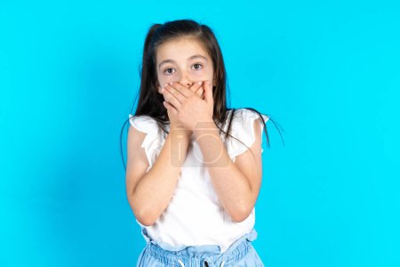 Photo for Stunned kid covers both hands on mouth, afraids of something astonishing. Beautiful caucasian little girl posing over blue studio background - Royalty Free Image