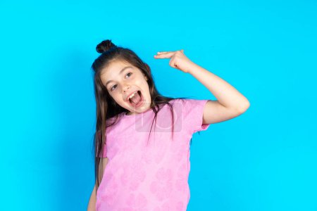 Photo for Kid foolishing around, shooting in temple with fingers, makes suicide gesture. Funny model makes finger gun pistol. Beautiful caucasian little girl posing over blue studio background - Royalty Free Image