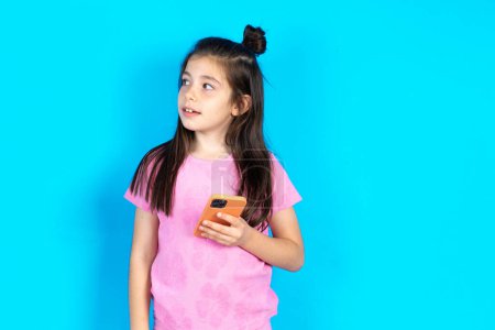 Photo for Kid holds mobile phone, uses high speed internet and social networks, concept of online communication and modern technologies. Beautiful caucasian little girl posing over blue studio background - Royalty Free Image