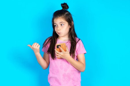 Photo for Kid points thumb away and shows blank space aside, holds mobile phone for sending text messages. Beautiful caucasian little girl posing over blue studio background - Royalty Free Image