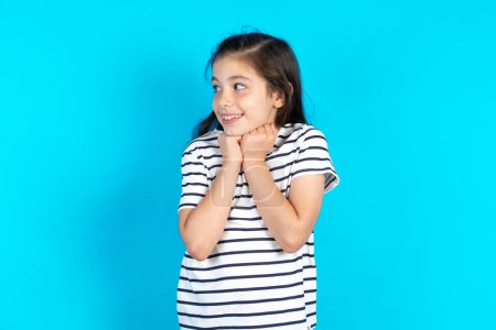 Photo for Happy kid anticipates something awesome happen, looks happily aside, keeps hands together near face, has glad expression. Beautiful caucasian girl posing over blue studio background - Royalty Free Image
