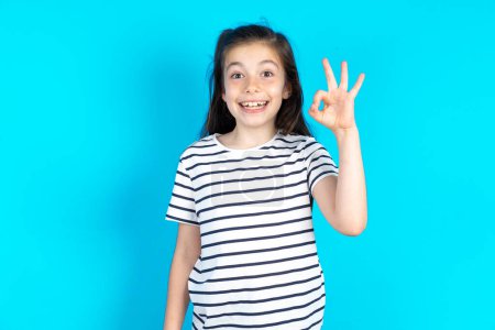 Foto de Feliz niña sonriente mostrando símbolo okey con su mano. Hermosa chica caucásica posando sobre fondo de estudio azul - Imagen libre de derechos