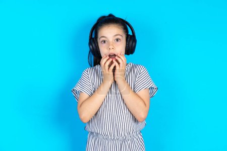 Photo for Shocked kid stares fearful at camera keeps mouth widely opened wears wireless stereo headphones on ears. Beautiful caucasian girl in striped dress standing over blue studio background - Royalty Free Image