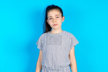 Photo for Displeased kid frowns face feels unhappy has some problems. Negative emotions and feelings concept.  Beautiful caucasian girl in striped t-shirt standing over blue studio background - Royalty Free Image