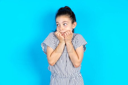 Photo for Terrified kid looks at empty space.  Beautiful caucasian girl in striped t-shirt standing over blue studio background - Royalty Free Image