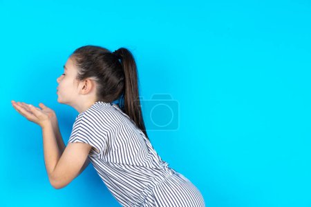 Téléchargez les photos : Vue latérale du profil, portrait d'un mignon enfant envoyant un baiser d'air. Belle fille caucasienne en t-shirt rayé debout sur fond de studio bleu - en image libre de droit