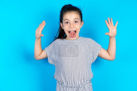 Photo for Delighted positive kid opens mouth and raising arms with palms up after having great result.  Beautiful caucasian girl in striped t-shirt standing over blue studio background - Royalty Free Image