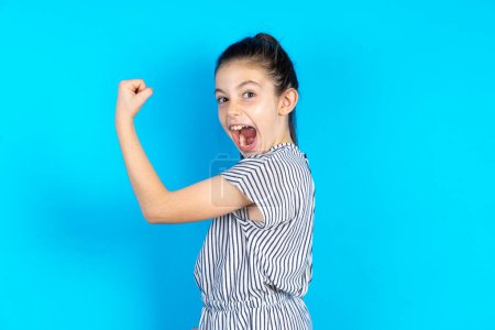 Photo for Profile side view portrait kid celebrates victory.  Beautiful caucasian girl in striped t-shirt standing over blue studio background - Royalty Free Image
