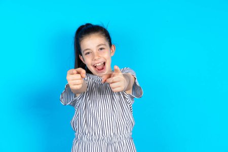 Photo for Beautiful smiling caucasian little girl in striped t-shirt standing over blue studio background poiting at camera - Royalty Free Image