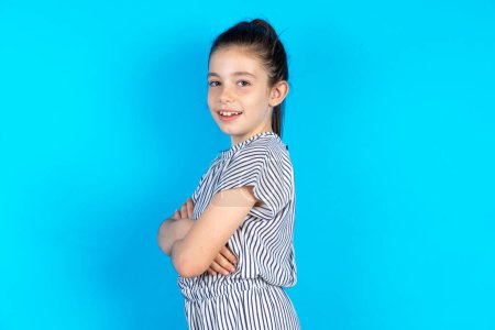 Téléchargez les photos : Portrait de belle petite fille caucasienne en t-shirt rayé debout sur fond de studio bleu avec bras croisés et sourire - en image libre de droit