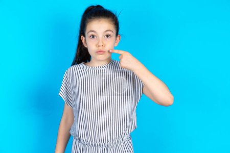 Photo for Charming kid pointing on pout lips with forefinger, showing effect after lifting procedure. Beautiful caucasian little girl in striped t-shirt standing over blue studio background - Royalty Free Image