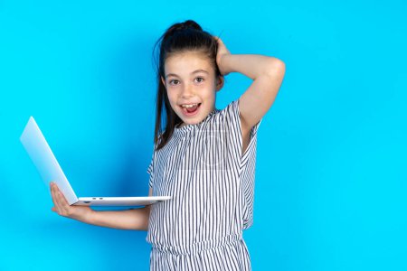 Photo for Photo of amazed Beautiful caucasian little girl in striped t-shirt standing over blue studio background holding modern gadget, arm on head - Royalty Free Image