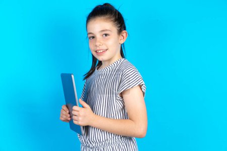 Téléchargez les photos : Photo optimiste Belle petite fille caucasienne en t-shirt rayé debout sur fond bleu studio tenant tablette pc - en image libre de droit