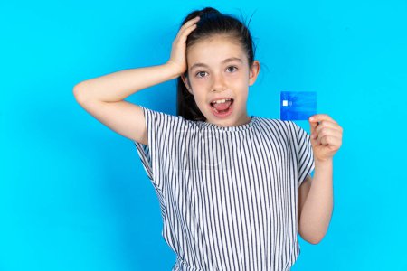 Téléchargez les photos : Impressionné drôle Belle fille enfant caucasien en t-shirt rayé debout sur fond de studio bleu avec la main sur la tête tenant la carte bancaire - en image libre de droit