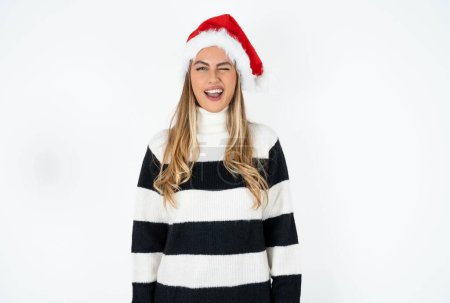 Foto de Coqueta joven caucásica con sombrero de Santa y suéter a rayas contra fondo blanco estudio sonriendo felizmente, parpadeando a la cámara de una manera lúdica, coqueteando con usted. - Imagen libre de derechos