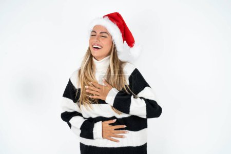 Foto de Joven mujer caucásica con sombrero de Santa y suéter a rayas contra fondo blanco estudio sonriendo y riendo en voz alta porque divertido broma loca con las manos en el cuerpo. - Imagen libre de derechos