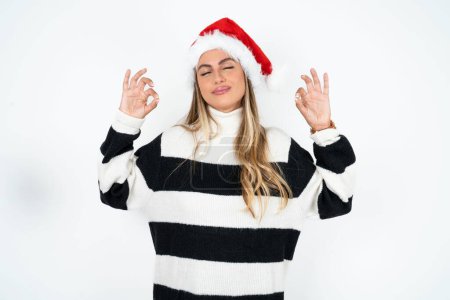 Téléchargez les photos : Jeune femme caucasienne portant le chapeau de Père Noël et pull rayé sur fond de studio blanc se détendre et sourire les yeux fermés faire geste de méditation avec les doigts. Concept de yoga. - en image libre de droit