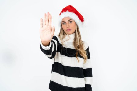 Téléchargez les photos : Jeune femme caucasienne portant le chapeau de Père Noël et pull rayé sur fond de studio blanc faire arrêter chanter avec la paume de la main. Expression d'avertissement avec geste négatif et sérieux sur le visage. - en image libre de droit