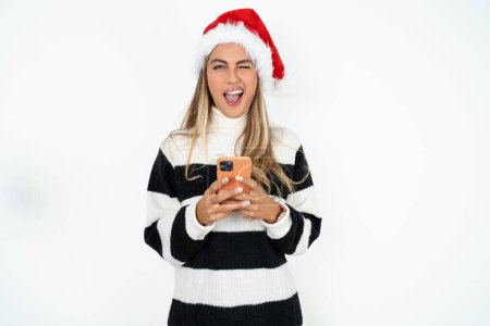 Téléchargez les photos : Jeune femme caucasienne excitée portant le chapeau de Père Noël et pull rayé sur fond blanc studio clin d'oeil et tenir les yeux utilisation de téléphone intelligent lire les nouvelles des réseaux sociaux - en image libre de droit