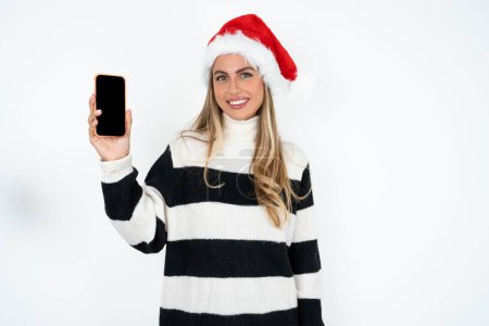 Photo for Smiling young caucasian woman wearing Santa hat and striped sweater against white studio background Mock up copy space. Hold mobile phone with blank empty screen - Royalty Free Image