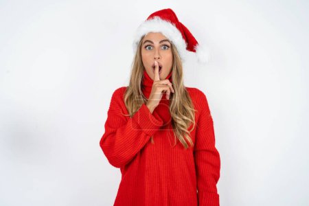 Photo for Surprised young caucasian woman wearing Santa hat and red sweater against white studio background makes silence gesture, keeps finger over lips and looks mysterious at camera - Royalty Free Image