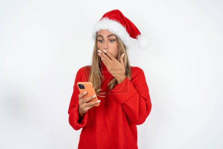 Photo for Young caucasian woman wearing Santa hat and red sweater against white studio background being deeply surprised, stares at smartphone display, reads shocking news on website, Omg, its horrible! - Royalty Free Image