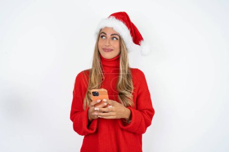 Photo for Young caucasian woman wearing Santa hat and red sweater against white studio background holds mobile phone uses high speed internet and social networks has online communication. Modern technologies concept - Royalty Free Image