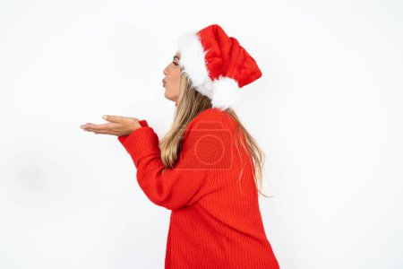 Téléchargez les photos : Profil vue de côté, portrait d'une jolie jeune femme caucasienne portant un chapeau de Père Noël et un pull rouge sur fond de studio blanc envoyant un baiser d'air - en image libre de droit