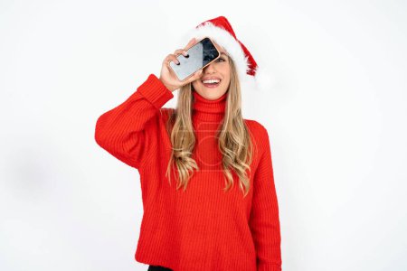 Foto de Joven mujer caucásica con sombrero de Santa y suéter rojo contra fondo de estudio blanco sosteniendo teléfono inteligente moderno que cubre un ojo mientras sonríe - Imagen libre de derechos