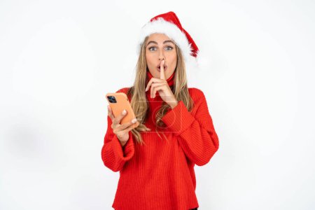 Photo for Young caucasian woman wearing Santa hat and red sweater against white studio background holding modern gadget ask not tell secrets - Royalty Free Image