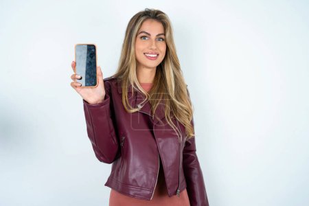 Téléchargez les photos : Souriant jeune femme caucasienne portant une veste en cuir tient le téléphone portable avec écran vide vierge - en image libre de droit