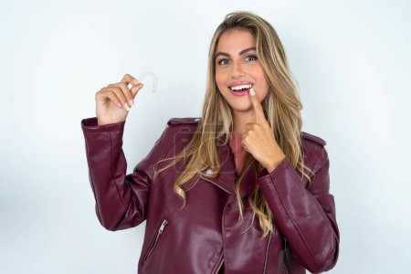 Photo for Young caucasian woman wearing leather jacket  holding an invisible aligner and pointing to her perfect straight teeth. Dental healthcare and confidence concept. - Royalty Free Image