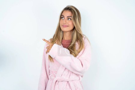 Photo for Young caucasian woman wearing bathrobe points at copy space and advertises something, advices best price. - Royalty Free Image