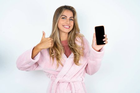 Foto de Joven mujer caucásica con albornoz Mostrando teléfono inteligente de pantalla en blanco, pulgar hacia arriba, recomienda nueva aplicación - Imagen libre de derechos
