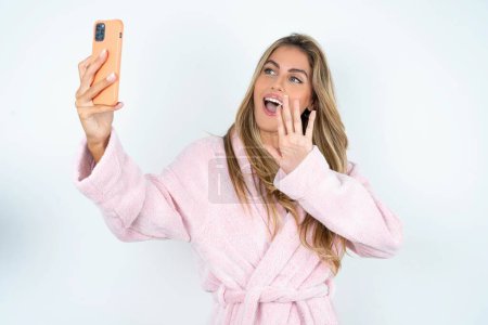Téléchargez les photos : Jeune femme caucasienne en peignoir tient téléphone portable moderne et fait ondes d'appel vidéo palme dans le geste bonjour. Personnes concept de technologie moderne - en image libre de droit