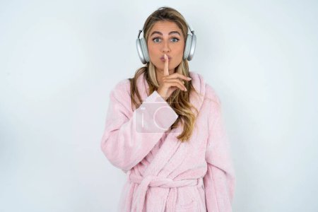 Photo for Young caucasian woman wearing bathrobe  making hush gesture with finger on her lips wearing  wireless headphones. Be quiet. - Royalty Free Image