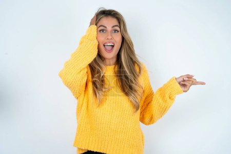 Photo for Surprised  young caucasian woman wearing yellow sweater pointing at empty space holding hand on head - Royalty Free Image
