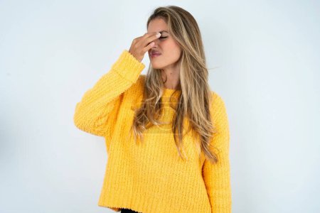 Foto de Mujer caucásica joven triste usando suéter amarillo que sufre de dolor de cabeza cogida de la mano en su cara - Imagen libre de derechos
