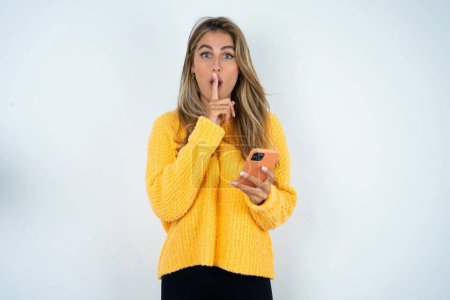 Téléchargez les photos : Jeune femme caucasienne portant un pull jaune tenant smartphone moderne demandant de ne pas dire de secrets - en image libre de droit