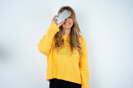 Téléchargez les photos : Femme caucasienne portant un pull jaune tenant smartphone moderne couvrant un oeil tout en souriant - en image libre de droit