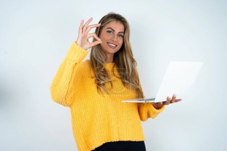 Téléchargez les photos : Positif jeune femme caucasienne portant un pull jaune détient netbook sans fil et montrant okey symbole - en image libre de droit