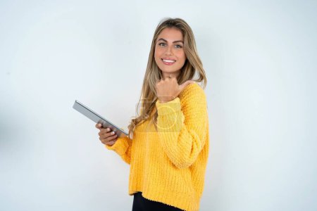 Téléchargez les photos : Jeune femme caucasienne portant pull jaune pointant vers l'espace vide tout en tenant ordinateur portable moderne - en image libre de droit