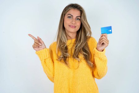 Téléchargez les photos : Souriant jeune femme caucasienne portant pull jaune montrant carte de débit pointant doigt espace vide - en image libre de droit