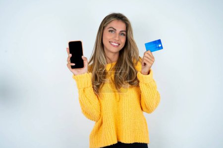 Téléchargez les photos : Jeune femme caucasienne portant un pull jaune ouvert compte bancaire, tenant smartphone et carte de crédit, souriant, recommande d'utiliser l'application de magasinage en ligne - en image libre de droit