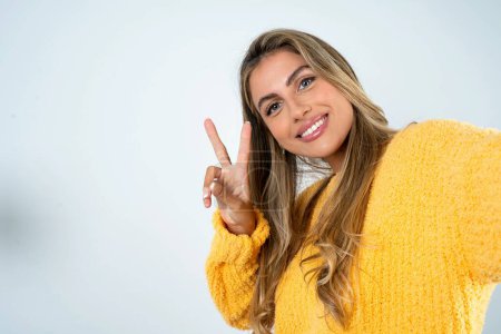 Téléchargez les photos : Positif jeune femme caucasienne portant pull jaune prenant selfie faisant v-signe - en image libre de droit