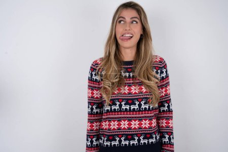 Photo for Funny caucasian woman wearing christmas sweater makes grimace and crosses eyes plays fool has fun alone sticks out tongue. - Royalty Free Image