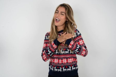 Téléchargez les photos : Femme caucasienne portant un pull de Noël exprime le bonheur, rit agréablement, garde les mains sur le cœur - en image libre de droit